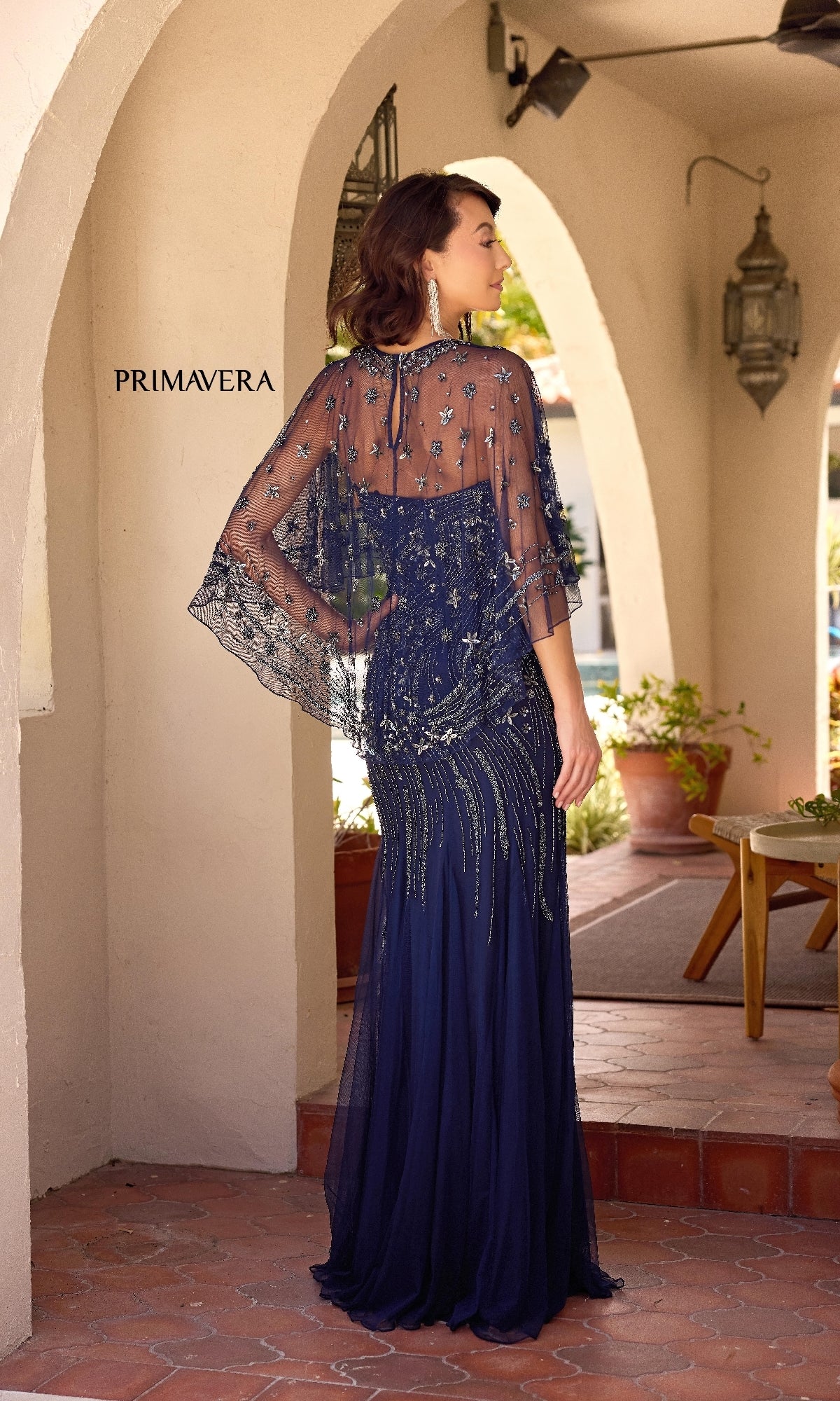 A woman stands on a covered patio wearing the Primavera 13145: a navy blue, strapless long formal dress with a mermaid skirt, intricate beadwork, and an elegant sheer removable capelet.